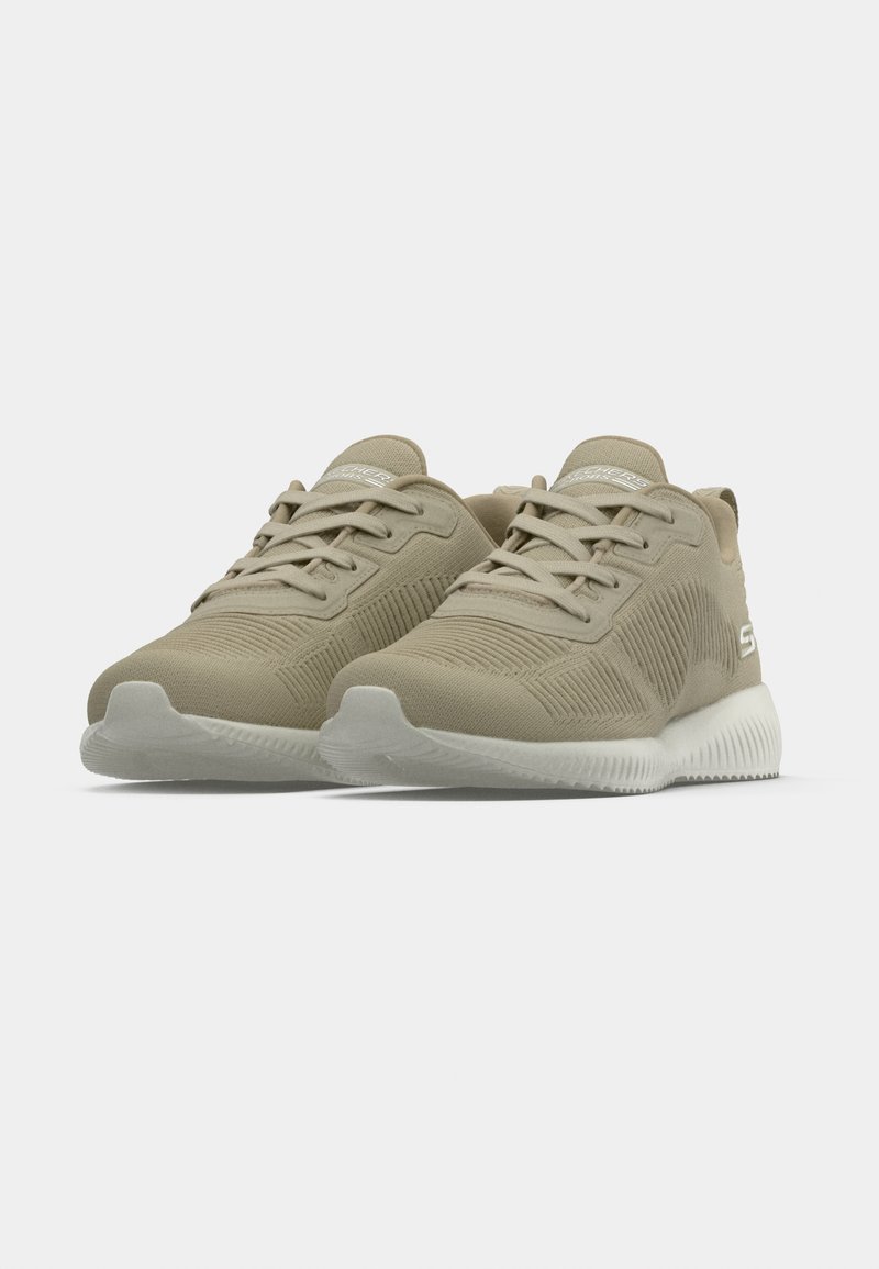 Zapatillas deportivas beige con parte superior texturizada, cierre de cordones y suela de goma blanca. Presenta una lengüeta lisa y un branding mínimo.