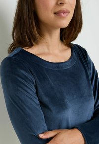 Top in velluto blu navy con scollo rotondo, maniche lunghe e trama liscia, caratterizzato da una lieve lucentezza e una vestibilità morbida.