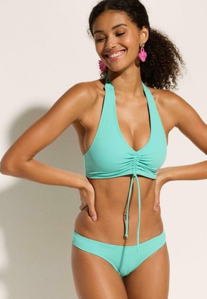 Donna sorridente con capelli ricci che indossa un bikini a halter turchese e orecchini rosa, posa con le mani sui fianchi su uno sfondo chiaro.