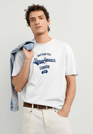 Junger Mann trägt ein weißes T-Shirt mit dem Aufdruck "Pepe Jeans London", hält eine Jeansjacke über der Schulter und steht vor einem schlichten Hintergrund.