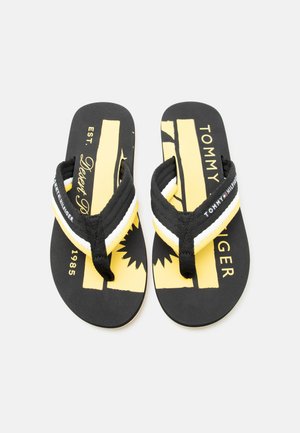 Paar schwarze Tommy Hilfiger Flip-Flops mit gelben Grafiksohlen und schwarzen, weißen sowie gelben Riemen, die den Markennamen und das Logo tragen.