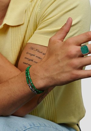 Grøn perlekæde med sølv detaljer, kraftig grøn stens ring, iført lysegul strikket polo, viser naturlig hudtekstur.