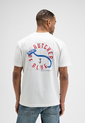 Man draagt een wit T-shirt met een blauwe haak en rode tekst "Butcher of Blue" op de rug, gecombineerd met blauwe spijkerbroek.