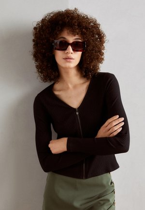 Black ribbed long-sleeve top with a zip closure, paired with a green skirt featuring button accents. Model has curly hair and sunglasses.