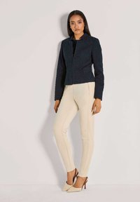 Marineblauwe cropped jas van gestructureerde stof, met hoge kraag en sluiting aan de voorkant, gecombineerd met crèmewitte slim-fit broek en beige hoge hakken.