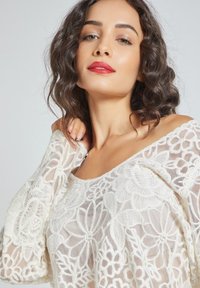 Top blanco transparente con intrincados patrones de encaje floral, diseño de hombros descubiertos, mangas largas y una silueta relajada.