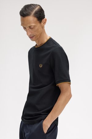 Uomo con camicia nera a maniche corte con rifiniture dorate ed emblema, in piedi con la testa inclinata verso il basso e le mani in tasca su sfondo semplice.