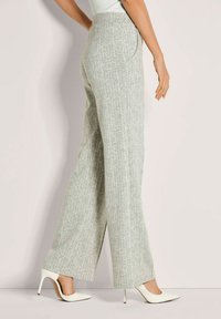 Vrouw in een lichtgrijze, wijde, hoge taille broek met een patroon en witte, spitse, hoge hakken, staand in profiel tegen een effen muur.