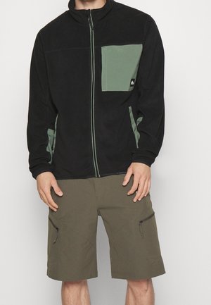 Uomo che indossa una giacca nera con cerniera e tasche verdi e pantaloncini cargo verde oliva con tasche con cerniera.