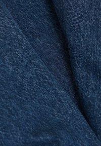 Primer plano de tela de mezclilla azul oscuro con textura de tejido visible y pliegues diagonales que crean sombras.