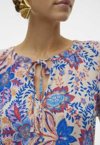 Blumenbluse mit beigem Hintergrund und blauen, rosa und orangefarbenen Mustern. Mit einer Bindeschließe am Hals und einem runden Ausschnitt.
