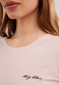 Šviesiai rožinė marškinėliai su apvaliu iškirpimu, trumpomis rankovėmis ir tamsiai mėlyna siuvinėta tekstu kairėje krūtinės pusėje. Minkšta, glotni audinio tekstūra.