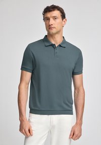 Dunkelblaugrünes Poloshirt mit kurzen Ärmeln, Drei-Knopf-Leiste und geripptem Kragen. Tragen Sie es mit weißen Hosen. Glatte Stofftextur.