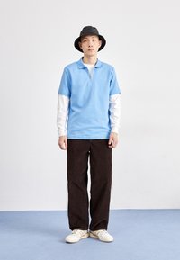 Polo azul claro com fecho e tecido texturizado, sobre uma camisa de manga longa branca. Calças de veludo cotelê castanhas e ténis bege. Chapéu bucket preto.