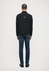 Camisa preta de manga longa com colarinho, apresentando um logotipo discreto nas costas. Combinada com jeans azuis e sapatos casuais pretos.