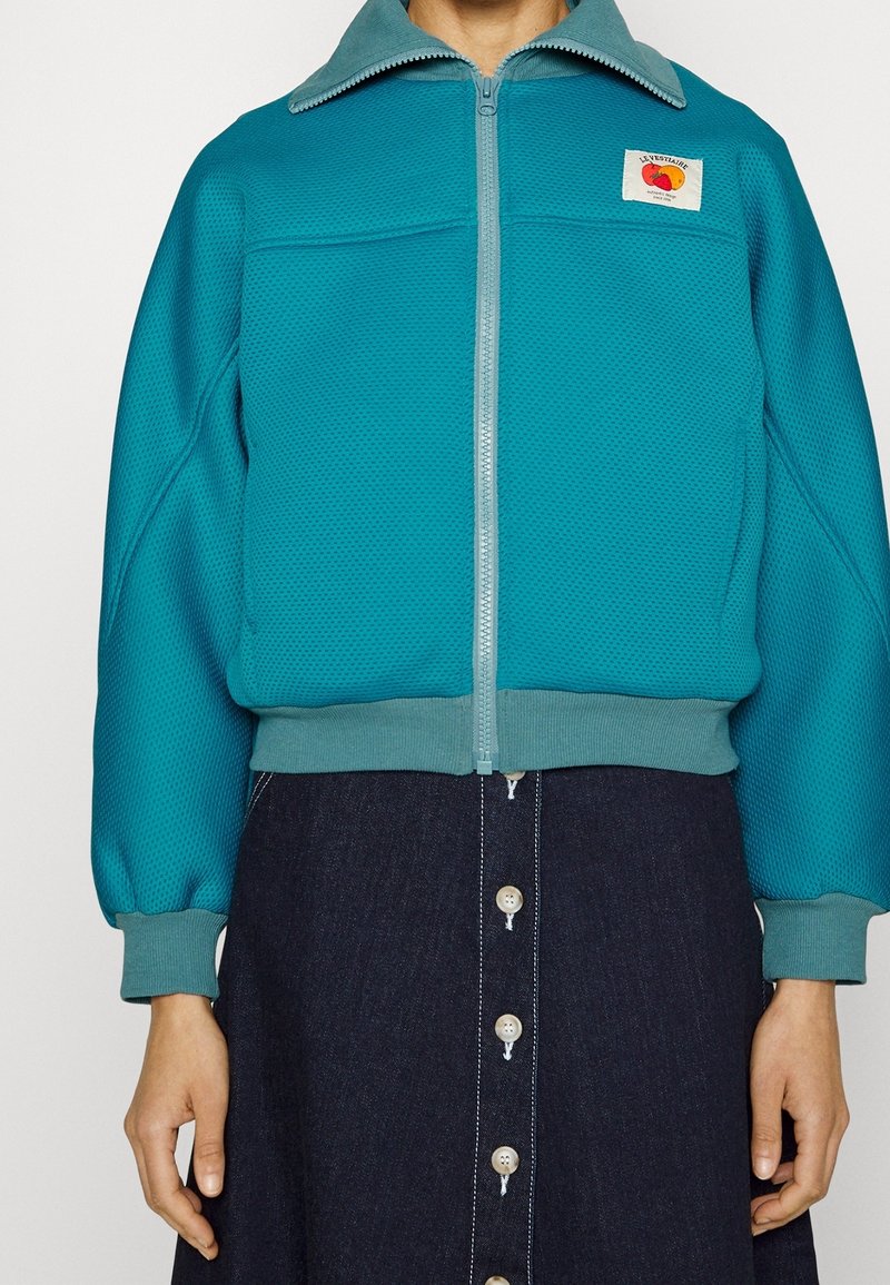 Turkos zip-up jacka i texturerad meshtyg, med kontrasterande ljusgrå krage och en patch-logga. Bärs över en jeanskjol med knappar.