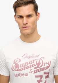 Hvid bomulds t-shirt med et falmet pink grafisk tekstdesign, der læser "Super Dry" med afrundede kanter og stiliserede accenter.