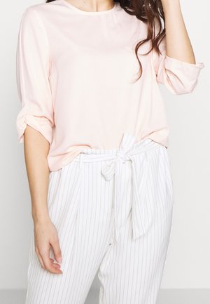 Kobieta ubrana w jasnoróżową bluzkę z długim rękawem, z podwiniętymi rękawami, oraz białe, wysoki stan spodnie w prążki, związane materiałowym paskiem.