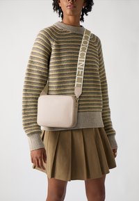 Borsa a tracolla in pelle beige con superficie testurizzata, forma rettangolare e una tracolla con branding ripetuto. Indossata con un maglione a righe e una gonna.