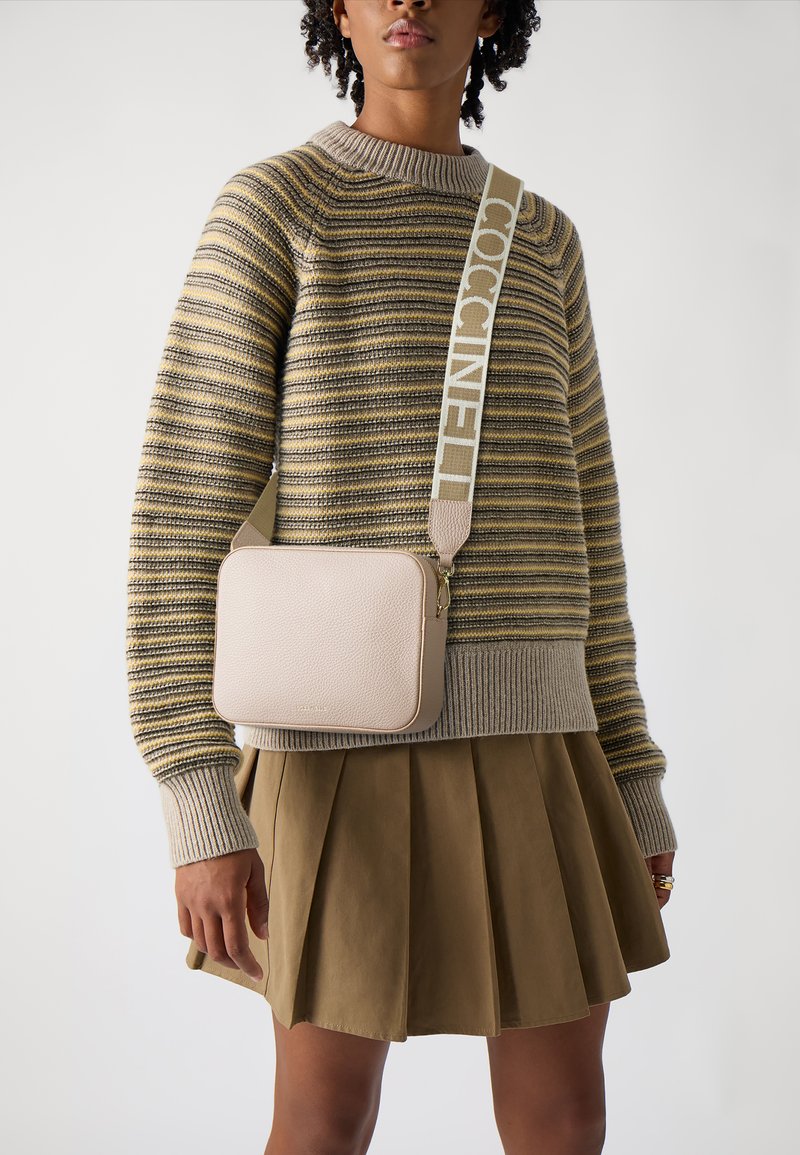 Borsa a tracolla in pelle beige con superficie testurizzata, forma rettangolare e una tracolla con branding ripetuto. Indossata con un maglione a righe e una gonna.