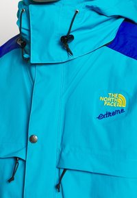 Jasnoniebieska wodoodporna kurtka z wysokim kołnierzem, regulowanymi troczkami i haftowanymi logo w żółtym i niebieskim kolorze. Kieszenie z zamkiem błyskawicznym z przodu.