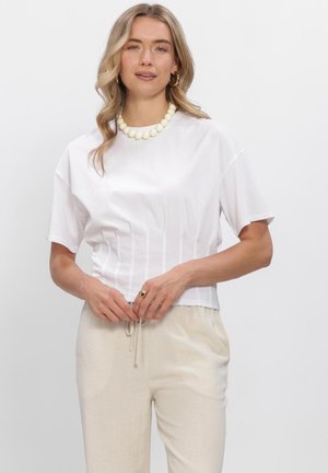 Vrouw met blond haar die een wit topje met korte mouwen draagt, beige broek met trekkoord, een witte kralenketting en een gouden ring, staand tegen een effen achtergrond.