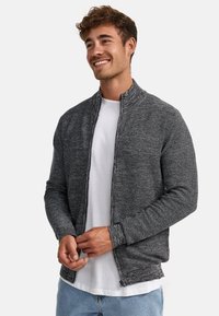 Cazadora gris con cremallera y acabado texturizado, que presenta un cuello alto y mangas largas, superpuesta sobre una camiseta blanca.