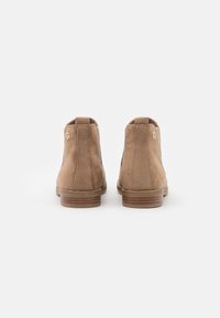 Botas de tobillo de ante en color tostado con punta redonda, tacón bajo y apilado, y un acento dorado. Presentan una textura suave y detalles de costura discretos.