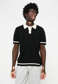 Fekete, rövid ujjú polo ing, krémszínű gallérral és csíkos díszítéssel. Világosszürke tréningnadrággal viselve. Pamut keverék anyagból készült, laza fazon.