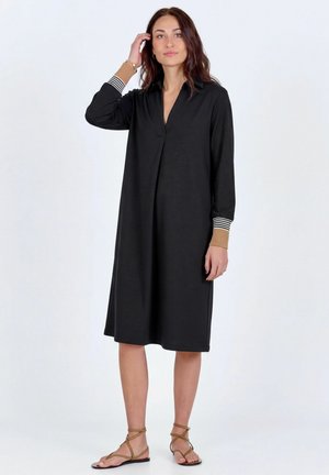 Žena v čiernych midi šatách s dlhým rukávom, ktoré majú hnedé a prúžkované manžety, a v hnedých sandáloch na plochom opätku s remienkami, stojaca pred jednoduchým pozadím.