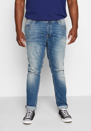 Homme portant un jean bleu délavé avec des revers roulés et des baskets montantes noires à lacets blancs, debout devant un fond uni.