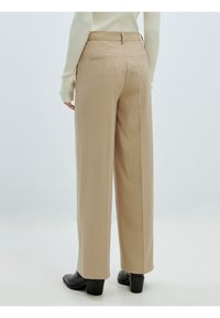 Beige gestreifte weit geschnittene Hose mit hoher Taille, versehen mit Gesäßtaschen und einer glatten Oberfläche. Kombiniert mit schwarzen Absatzstiefeln.