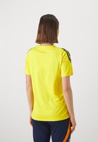 Žlutý sportovní dres s kulatým výstřihem, krátkými rukávy a černými profilovými pruhy na ramenou. Pohodlná, lehká tkanina.
