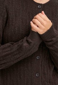 Ruskea teksturoitu neulecardigan, jossa on ribbaatut hihansuut ja nappilista. Sisältää kolme tummanväristä nappia ja pehmeää, pörröistä kangasta.