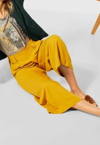 Gula vida byxor i slätt tyg med framveck. Bärs med en grå grafisk t-shirt och bruna sandaler.