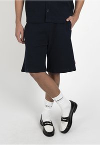 Personne portant un short noir, des chaussettes hautes blanches avec du texte et des mocassins noir et blanc, debout avec une jambe croisée sur l'autre.