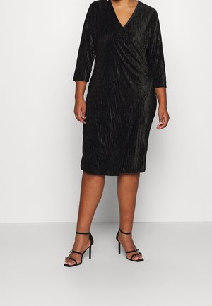 Femme portant une robe en velours noire longueur genou avec manches trois-quarts et des sandales noires à talons hauts avec brides à la cheville.