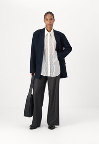 Marineblå overdimensjonert blazer, hvit blonde skjorte med knappestolpe, grå vide bukser, svart veske og svarte sko med dekorative detaljer.