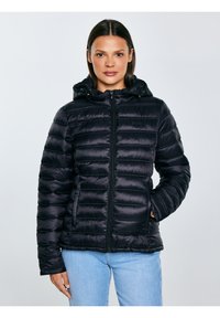 Sieviete, tērpusies melnā kostītā cimdu kapucēm apskautā jakā, aizpogāta, ar vienu roku kabatā, valkājot gaiši zilas džinsa bikses, pret vienkāršu fonu.