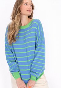 Blau- und grün gestreifter Pullover mit einem grünen gerippten Halsausschnitt und Bündchen. Weiche Textur; lockere Passform; lange Ärmel; überschnittene Schulternähte.
