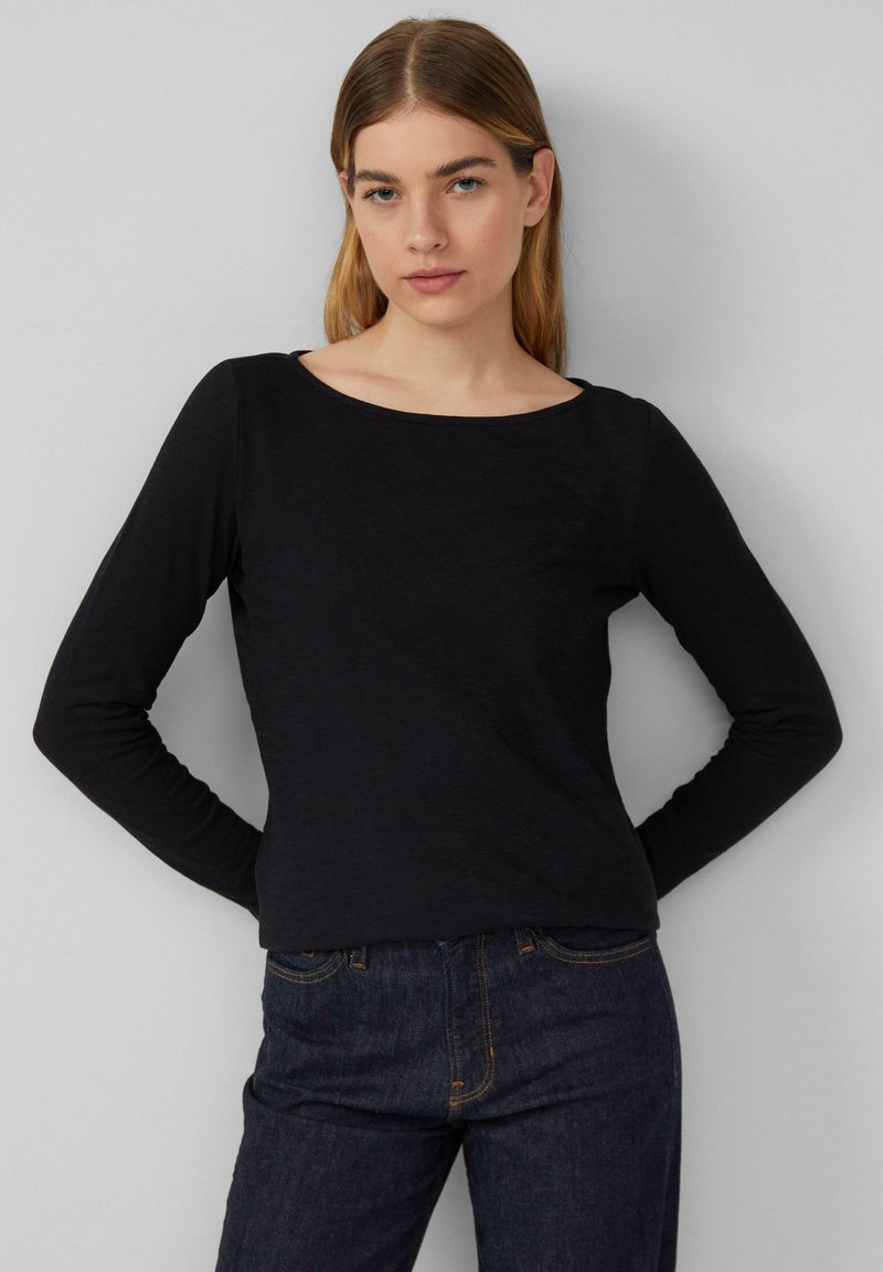 Schwarzes Langarmoberteil mit rundem Ausschnitt, aus weichem Stoff gefertigt. Kombiniert mit dunklen Jeans mit gelben Stichdetails.