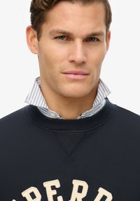 Sudadera navy con texto beige bordado, superpuesta a una camisa de collar a rayas azul y blanca. Tejido suave y escote estructurado.