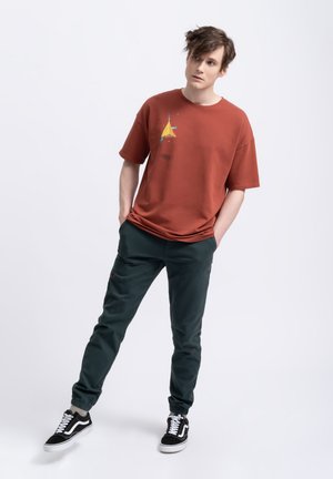 Roestkleurig oversized t-shirt met abstract ontwerp in geel en blauw op de borst, gecombineerd met donkergroene omgeslagen broek en zwarte sneakers.