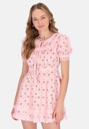 Ung kvinde med langt bølget hår iført en lyserød blomsterkjole med puffede ærmer, flæsedetaljer og bindebånd ved halsudskæringen på en hvid baggrund.