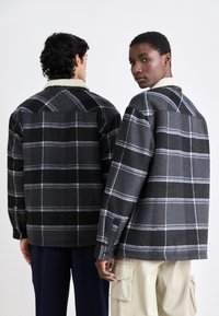 Zwei Personen tragen schwarze und graue karierte Flanelljacken mit einem weichen, weißen Kragen. Die Jacken haben eine lockere Passform und geknöpfte Manschetten.