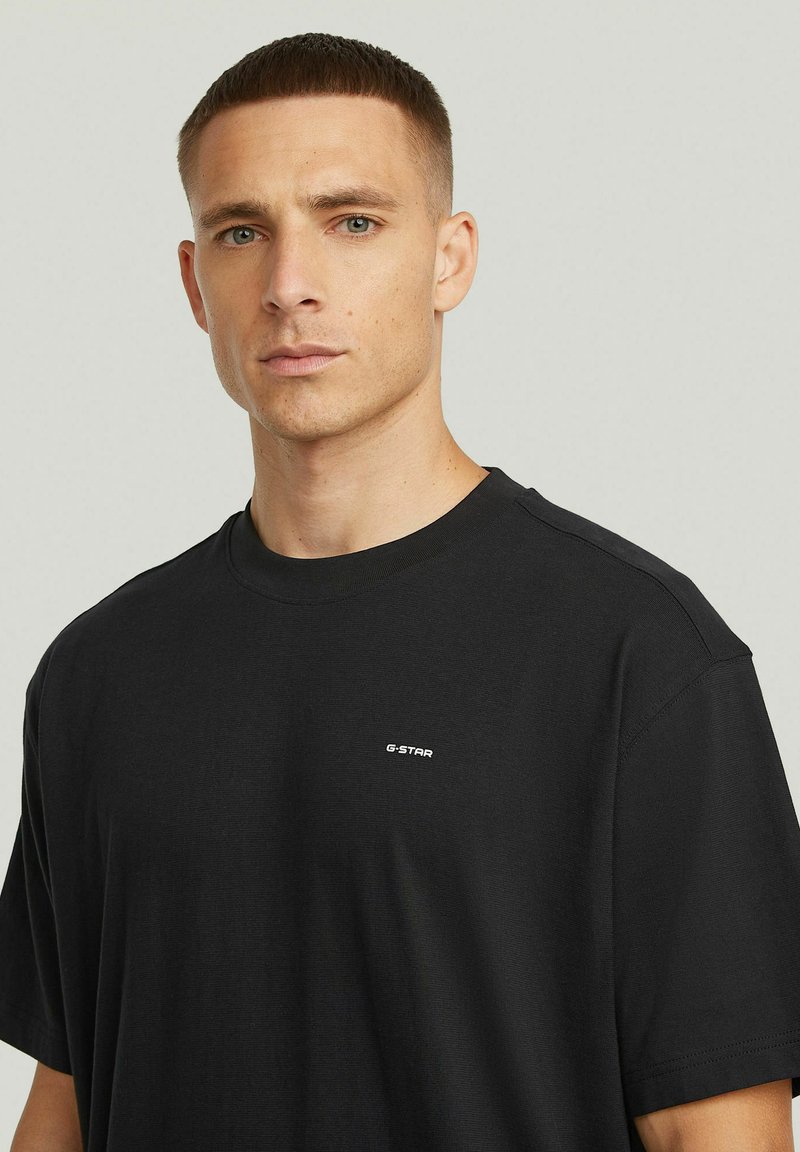 Uomo con capelli corti che indossa una maglietta nera semplice di G-Star, rivolto verso l'obiettivo su uno sfondo grigio chiaro.