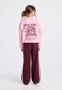 Femme avec des cheveux bruns en chignon portant un sweat-shirt rose avec un imprimé de boulangerie de bagels de New York et un pantalon bordeaux avec des rayures blanches sur les côtés.