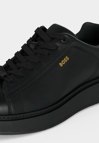 Μαύρο δερμάτινο sneaker χαμηλού ύψους με χοντρή σόλα, μαύρα κορδόνια και χρυσό λογότυπο "BOSS" στα πλευρά.