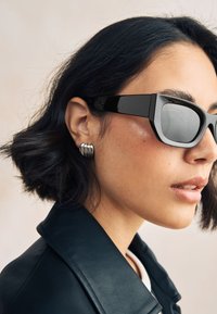 Schwarze rechteckige Sonnenbrille mit glänzendem Rahmen, getragen zu einer schwarzen Lederjacke und silbernen Ohrringen. Weich strukturierte Haare umrahmen das Gesicht.