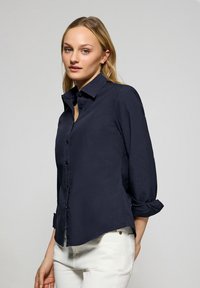 Marineblauwe blouse met knopen en een puntige kraag, lange mouwen opgerold bij de manchet en een licht gebogen zoom. Soepele stofstructuur.